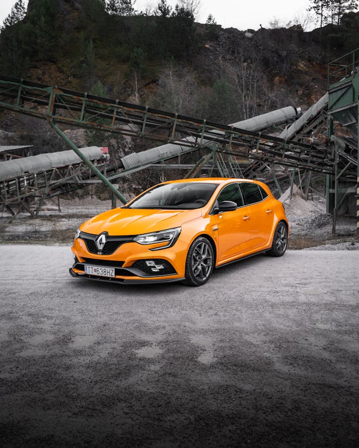 A vibrant orange sports car parked in an industrial area with machinery.